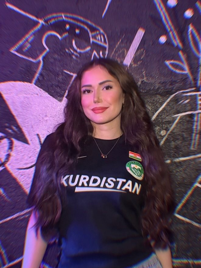 Jeune femme kurde en maillot Kurdistan Nike noir devant une fresque murale